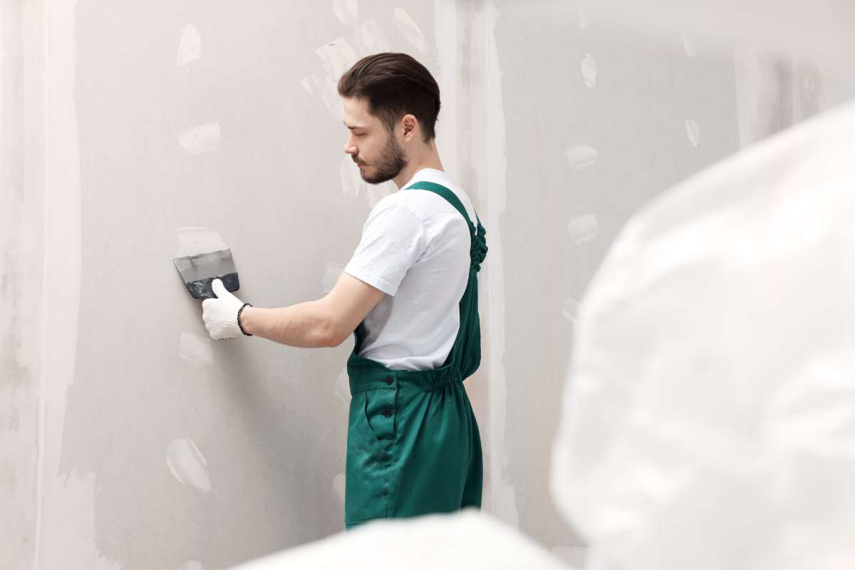 Worker in uniform plastering wall with putty knife indoors