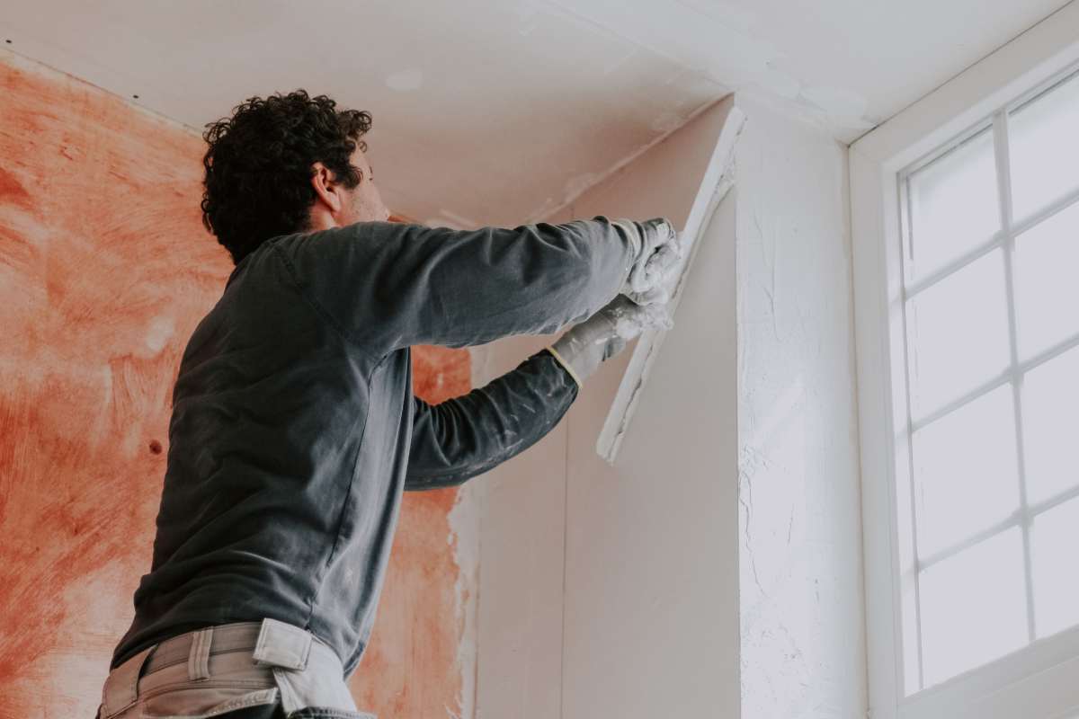 One young Caucasian curly-haired male plasterer in a uniform and construction gloves pulls off a large spatula and putty from the wall near the window, standing on a stepladder, lower side close-up view.
